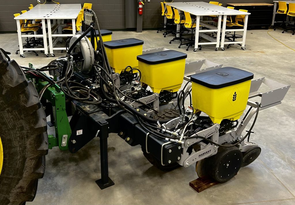Harvest International planter attached to the back of a tractor. There are four yellow tubs, evenly spaced, and a metal tray with open end behind the tubs.