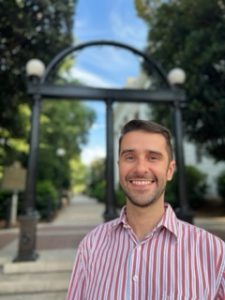 Leo Bastos standing in front of the UGA arch, wearing a red and white striped button up shirt.