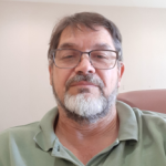 Headshot of Glen Rains, who is wearing a pale green collared polo shirt. He is wearing glasses and sitting in front of a white wall.