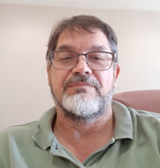 Headshot of Glen Rains, who is wearing a pale green collared polo shirt. He is wearing glasses and sitting in front of a white wall.