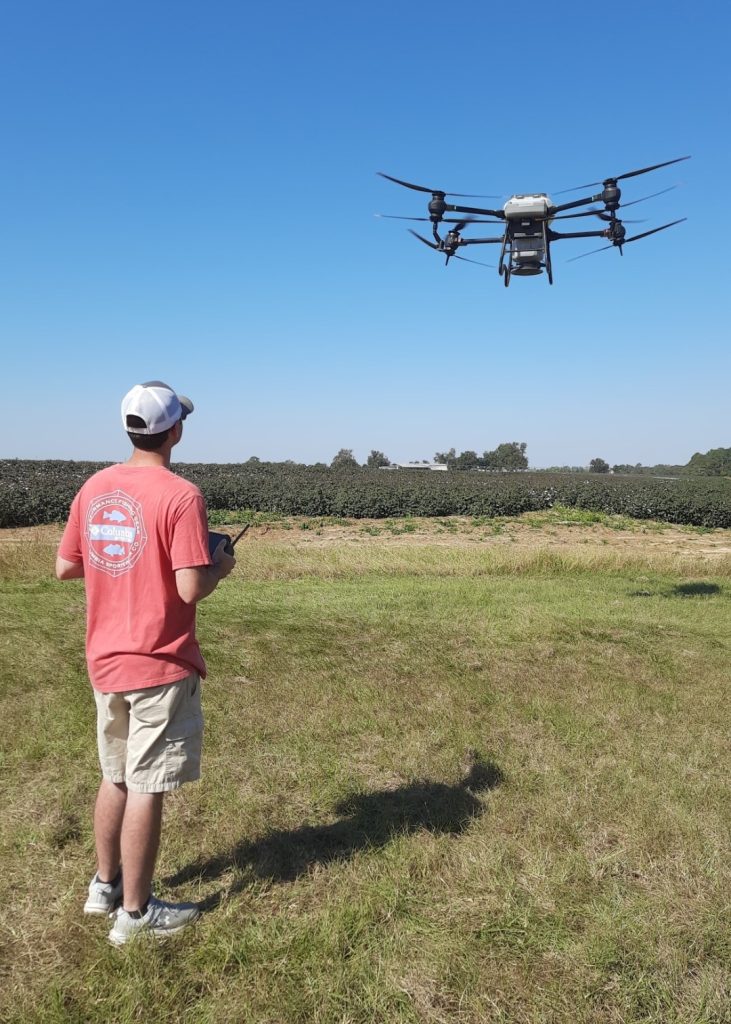 A person in a red shirt and khaki shorts is standing, back facing the camera, piloting a drone.
