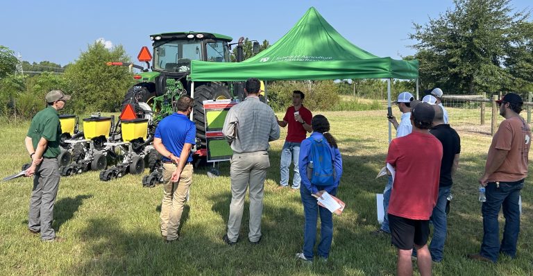 A green tent-topper is set up beside a planter (tractor). A man under neat the tent is talking and a group of people are gathered around, listening.