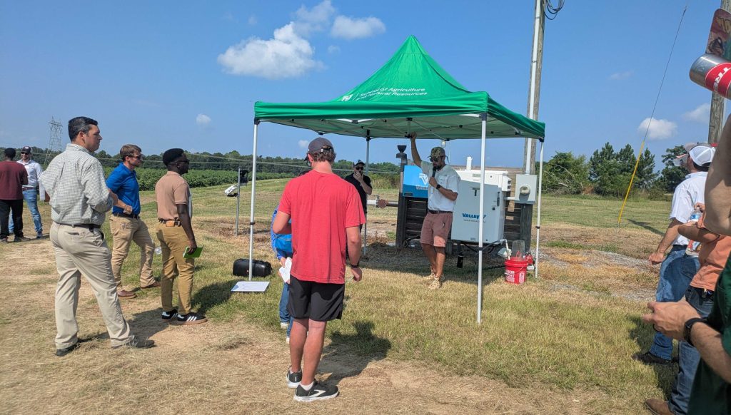 A green tent-topper is set up near an agricultural field. A man stands under it, talking to a small group of people gathered around.