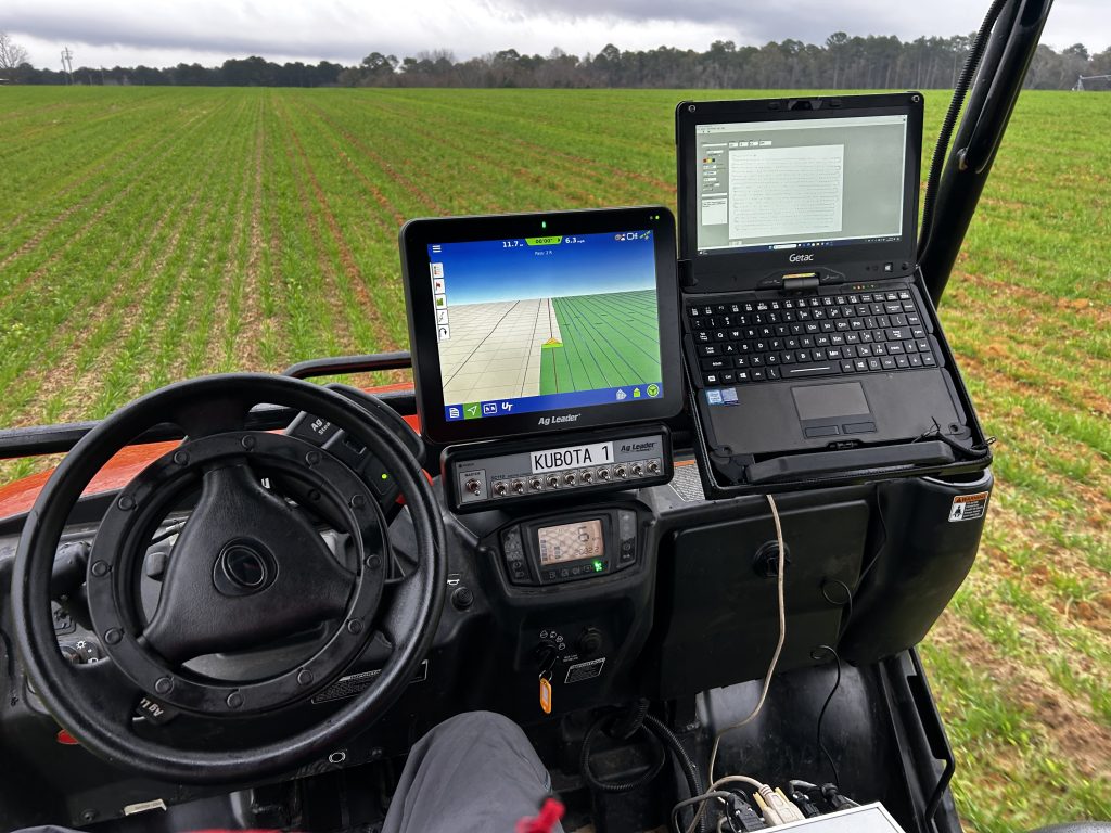 Photograph taken from the Kubota utility vehicle's driver seat. Two monitors are visible: one showing field guidance from AgLeader, the other a laptop receiving data from the Veris implement.