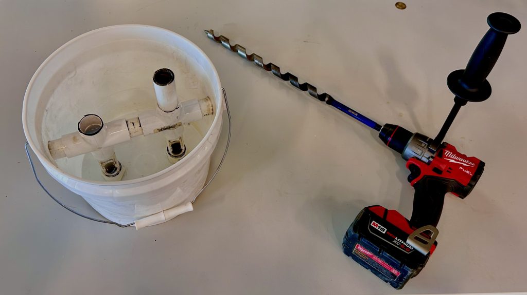 5 gallon bucket with PCV piping inside. An electric drill with an auger bit is beside the bucket.